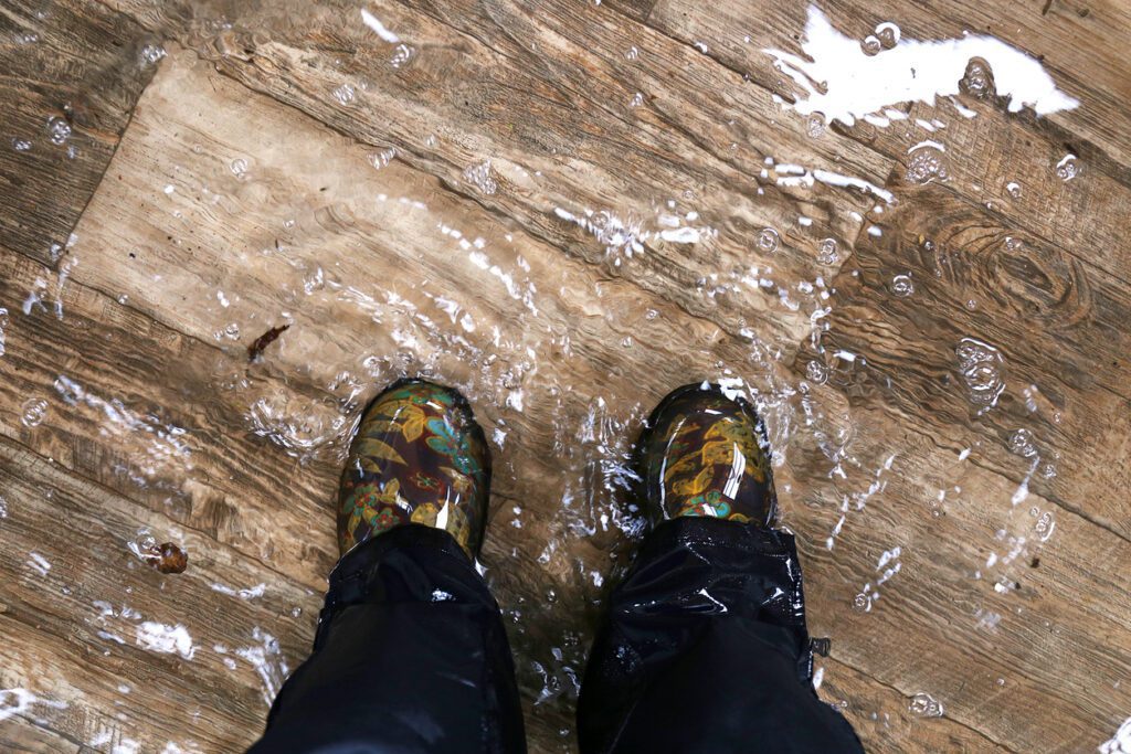 A close-up of a person standing in several inches of water, wearing waterproof boots, on a water-damaged wooden floor. The image highlights the flooding and damage to the floor caused by water.